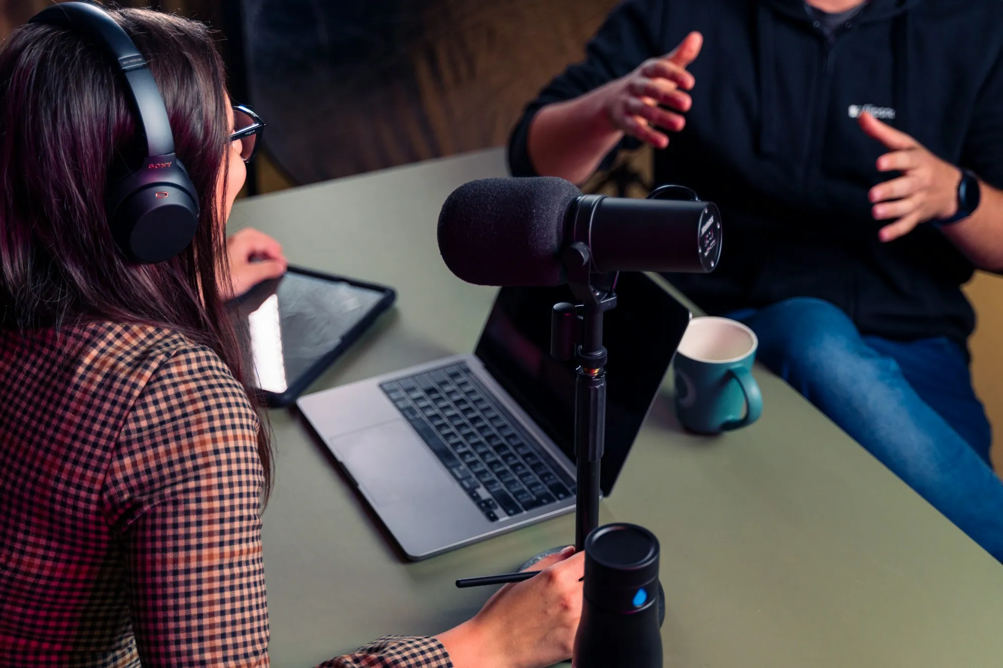 Two people in conversation around a podcast microphone, one wearing headphones at a laptop.